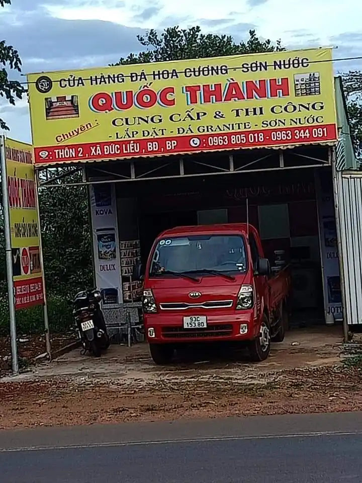 Xe tải giao hàng màu đỏ đậu dưới một biển hiệu doanh nghiệp màu vàng được viết bằng tiếng Việt.